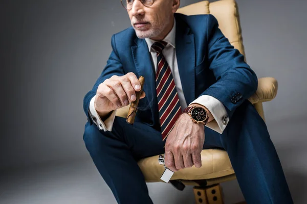 Reifer Mann im Anzug mit Zigarre und Feuerzeug im Sessel auf grau — Stockfoto