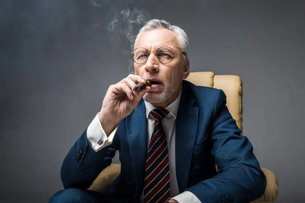 Mature businessman in suit and glasses smoking cigar on grey — Stock Photo