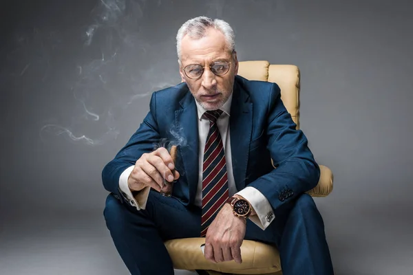 Pensive mature businessman in suit and glasses holding cigar while sitting in armchair on grey — Stock Photo