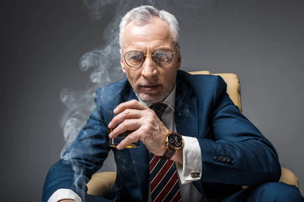 Mature businessman in suit and glasses holding glass of whiskey while sitting in armchair on grey with smoke — Stock Photo