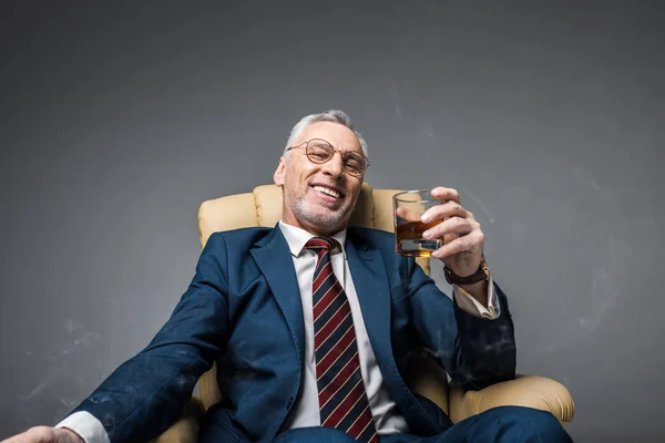 Cheerful mature businessman in suit holding glass of whiskey while sitting in armchair on grey — Stock Photo