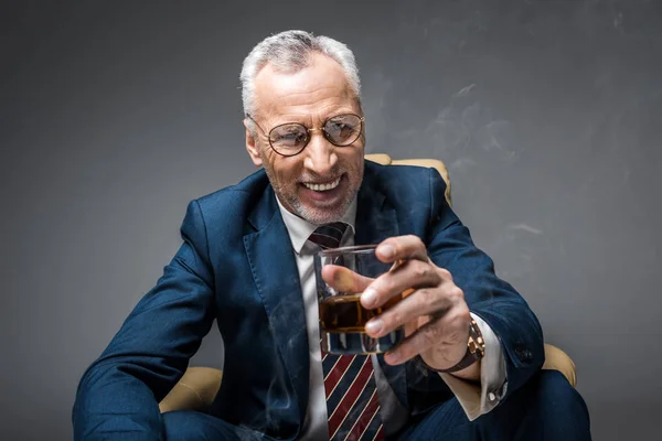 Счастливый зрелый бизнесмен в костюме держит стакан виски, сидя в кресле на сером — стоковое фото