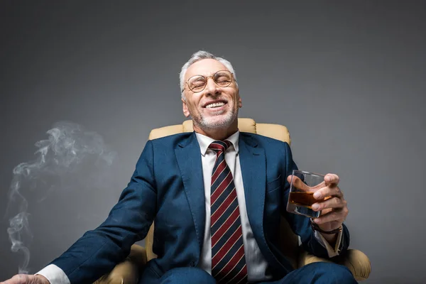 Sonriente hombre de negocios maduro en traje sosteniendo vaso de whisky mientras está sentado en sillón en gris - foto de stock