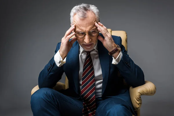 Stanco uomo d'affari maturo in giacca e cravatta avendo mal di testa mentre seduto in poltrona su grigio — Foto stock