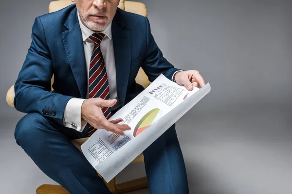 Обрізаний вид зрілого бізнесмена в костюмі, що тримає карти і графіки, сидячи в кріслі на сірому — стокове фото
