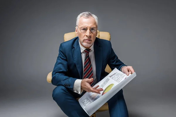 Mature businessman in suit and glasses holding charts and graphs while sitting in armchair on grey — Stock Photo