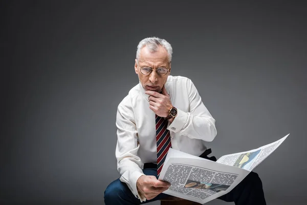 Pensive mature businessman in suit holding newspaper on grey — Stock Photo