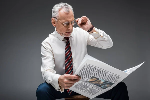 Mature businessman touching glasses while reading newspaper on grey — Stock Photo