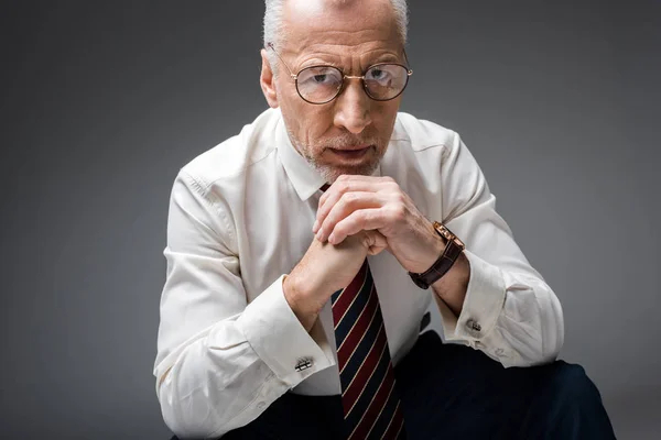 Hombre de negocios maduro en gafas y traje mirando a la cámara con las manos cerradas en gris - foto de stock