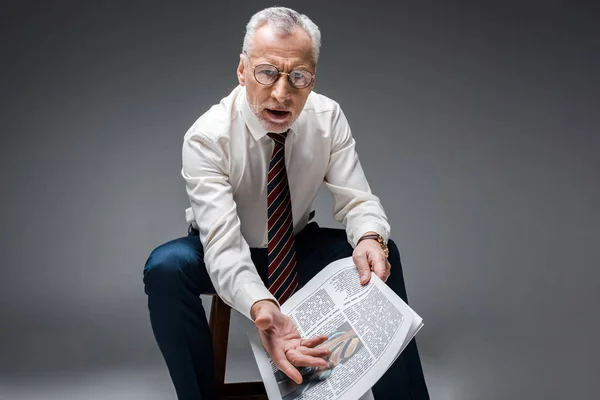 Mature businessman gesturing while holding newspaper on grey — Stock Photo