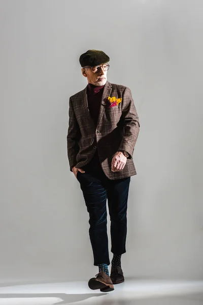 Hombre de mediana edad de moda en gafas y gorra caminando con la mano en el bolsillo en gris - foto de stock