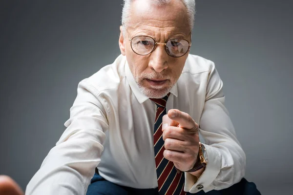 Homme d'affaires d'âge moyen dans des lunettes pointant du doigt tout en regardant la caméra sur gris — Photo de stock