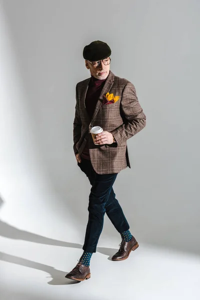 Elegante uomo di mezza età in occhiali e cappuccio camminare con tazza di carta su grigio — Foto stock