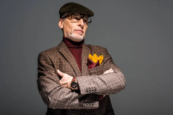 Stylish middle aged man in glasses and cap standing with crossed arms on grey — Stock Photo