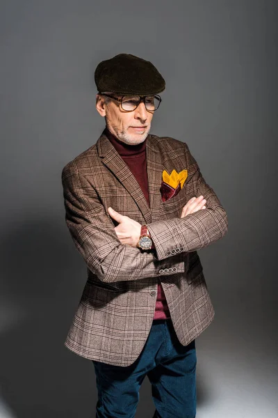 Homme d'âge moyen à la mode dans des lunettes et casquette debout avec les bras croisés sur gris — Photo de stock