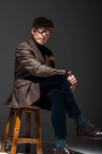 Stylish mature businessman in cap and glasses looking at camera on grey — Stock Photo