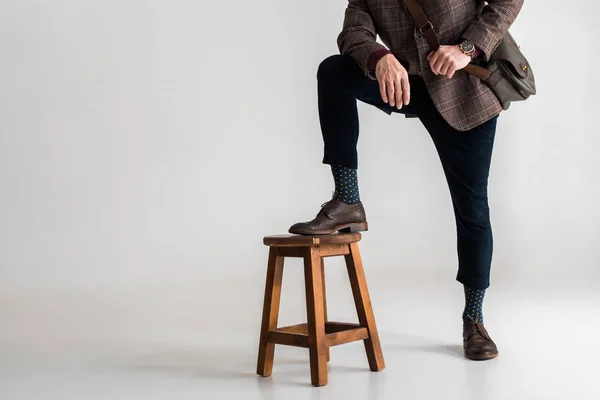 Ausgeschnittene Ansicht eines stilvollen reifen Mannes, der Bein auf Stuhl legt, während er auf grau steht — Stockfoto