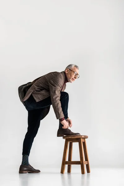 Trendy mature man putting leg on chair while tying shoelaces on grey — Stock Photo