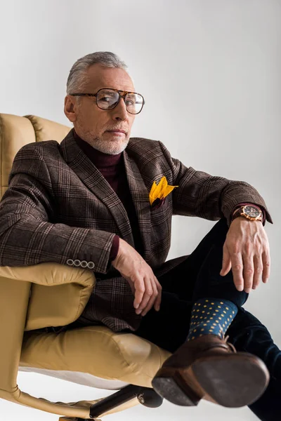Enfoque selectivo de hombre maduro de moda en gafas sentadas en sillón con piernas cruzadas aisladas en gris - foto de stock