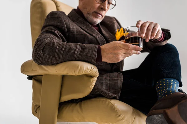 Messa a fuoco selettiva di uomo maturo alla moda seduto in poltrona con gambe incrociate e tenendo un bicchiere di whisky isolato sul grigio — Foto stock