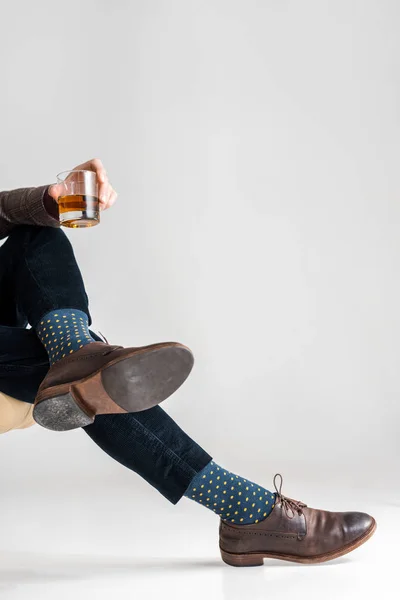 Abgeschnittene Ansicht eines reifen Mannes mit überkreuzten Beinen, der ein Glas Whiskey auf grau hält — Stockfoto