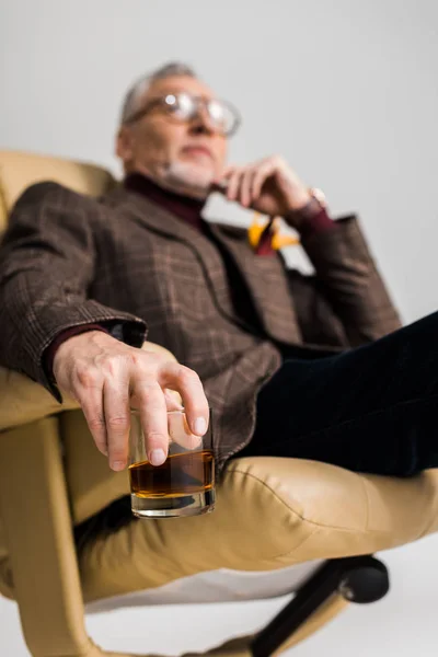 Selective focus of glass with whiskey in hand of mature man in glasses sitting in armchair isolated on grey — Stock Photo