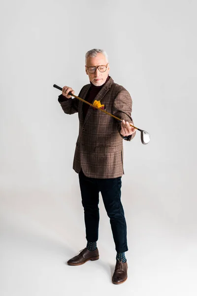 Stylish mature man in glasses holding golf club and while standing on grey — Stock Photo
