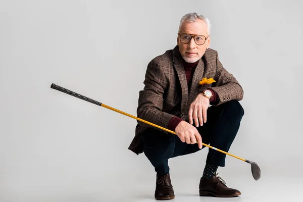 Hombre maduro de moda en gafas que sostienen el club de golf y mientras está sentado en gris - foto de stock