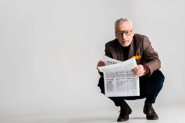 Trendy mature man in glasses holding newspaper while sitting on grey — Stock Photo