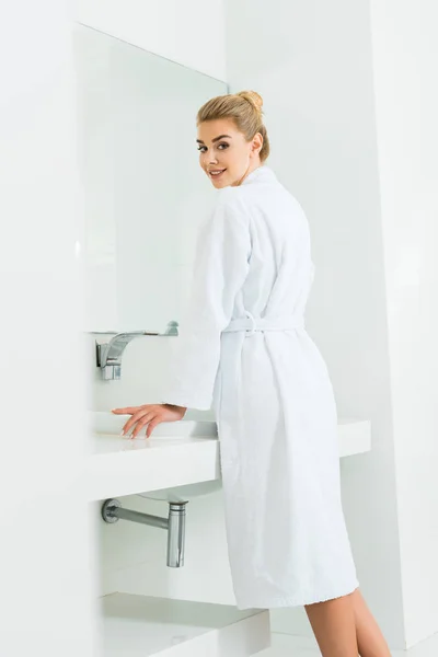Красивая и улыбающаяся женщина в белом халате смотрит на камеру в ванной комнате — стоковое фото