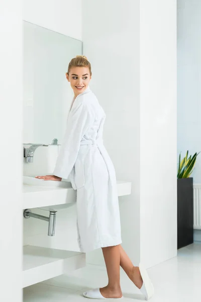 Hermosa y sonriente mujer en albornoz blanco y zapatillas mirando hacia otro lado en el baño - foto de stock