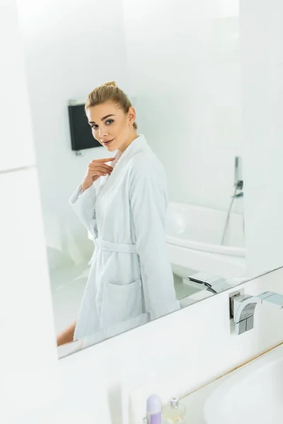 Красивая и улыбающаяся женщина в белом халате смотрит на зеркало в ванной комнате — стоковое фото
