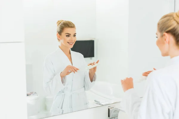 Selective focus of smiling woman in bathrobe applying toothpaste on toothbrush in bathroom — Stock Photo