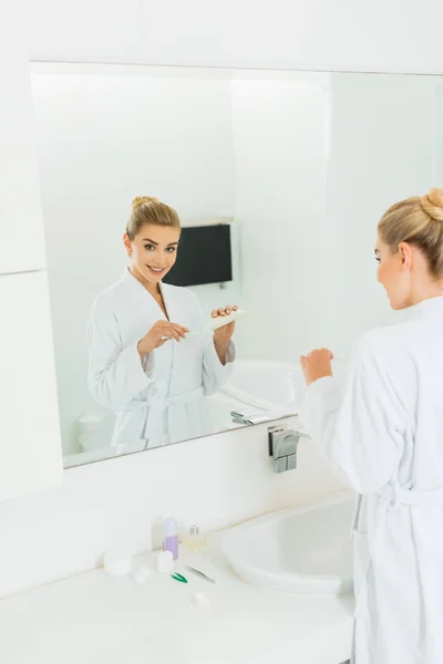 Женщина в халате наносит зубную пасту на зубную щетку и смотрит в зеркало — стоковое фото