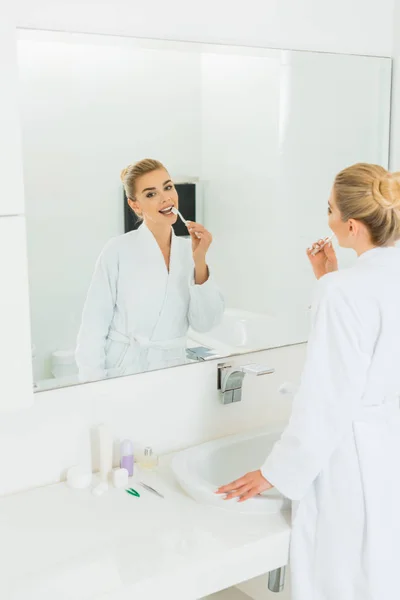 Beautiful woman in white bathrobe brushing teeth and looking at mirror — Stock Photo