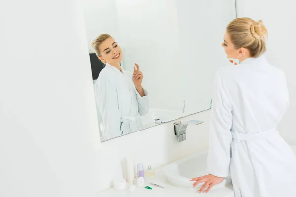 Selective focus of woman in white bathrobe holding toothbrush and looking at mirror — Stock Photo