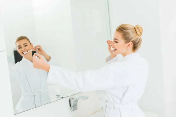 Selective focus of beautiful woman in white bathrobe brushing teeth and taking selfie — Stock Photo