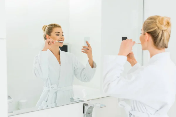 Selective focus of beautiful woman in white bathrobe brushing teeth and taking selfie in bathroom — Stock Photo
