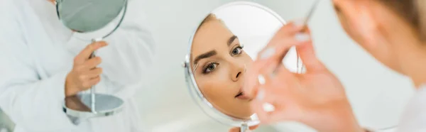 Panoramic shot of beautiful woman plucking eyebrows with tweezers in bathroom — Stock Photo