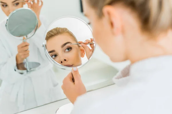 Selective focus of attractive woman plucking eyebrows with tweezers in bathroom — Stock Photo