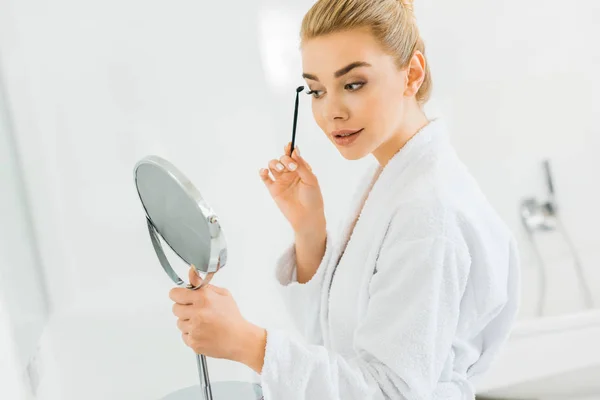 Attraktive Frau im weißen Bademantel, die Augenbrauen putzt und in den Spiegel schaut — Stockfoto