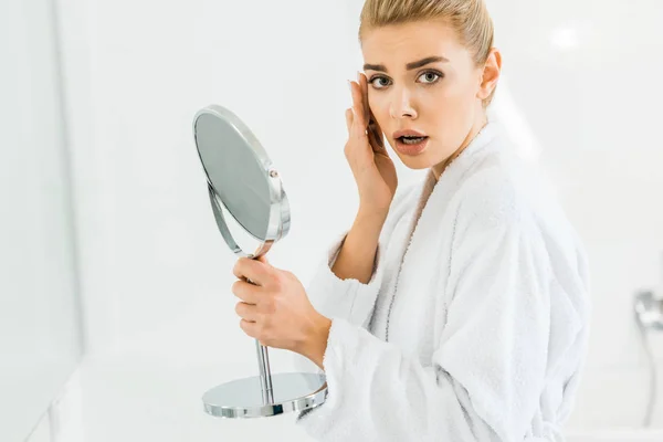 Überraschte und schöne Frau im weißen Bademantel mit Spiegel und Blick in die Kamera — Stockfoto