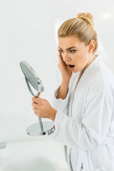 Шокированная и красивая женщина в белом халате смотрит на зеркало в ванной комнате — стоковое фото