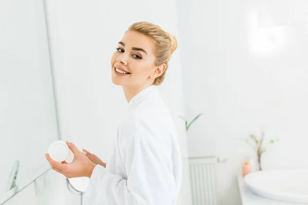 Mujer sonriente en albornoz blanco sosteniendo crema cosmética y mirando a la cámara en el baño - foto de stock