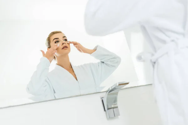Foco seletivo da mulher em roupão de banho branco aplicando creme facial no banheiro — Fotografia de Stock