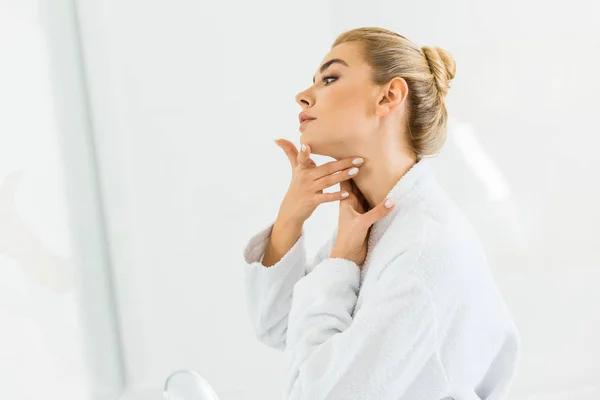 Attractive and blonde woman in white bathrobe applying face cream in bathroom — Stock Photo