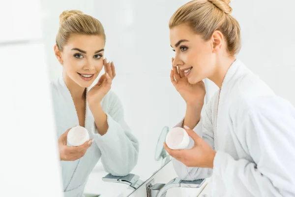 Foco seletivo de mulher atraente e loira em roupão branco aplicando creme facial e olhando para o espelho — Fotografia de Stock