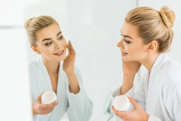 Messa a fuoco selettiva di attraente e sorridente donna in accappatoio bianco applicare la crema viso in bagno — Foto stock