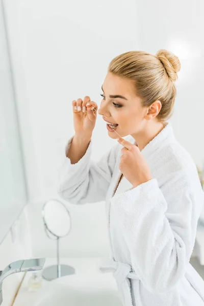Mujer atractiva y rubia en albornoz blanco cepillarse los dientes con hilo dental - foto de stock