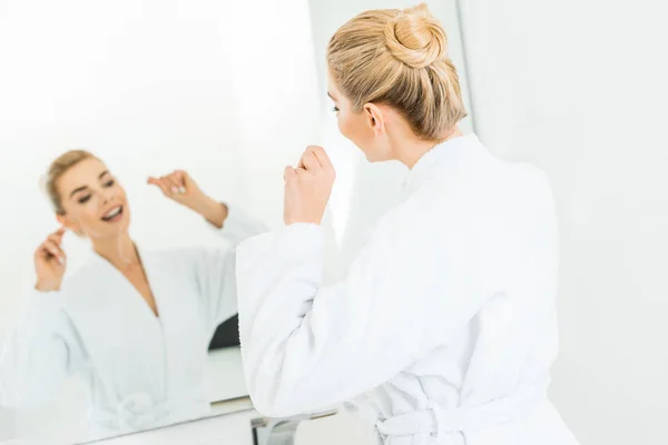 Selective focus of beautiful and blonde woman in white bathrobe brushing teeth with dental floss — Stock Photo